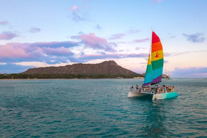Sailboat with rainbow sail on ocean with mountain and sky in background.