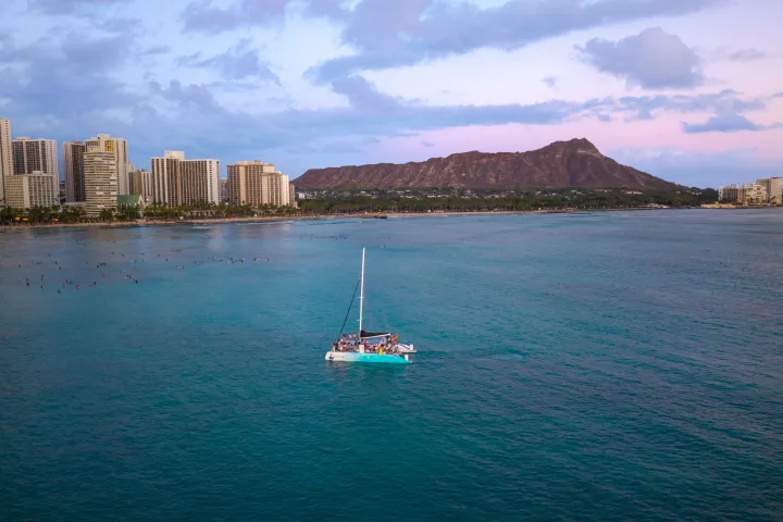 Sailboat on ocean with city skyline and mountain in the background during sunset.