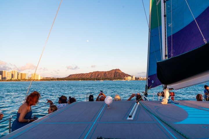 People on a sailboat with a view of a mountain and city skyline at sunset.