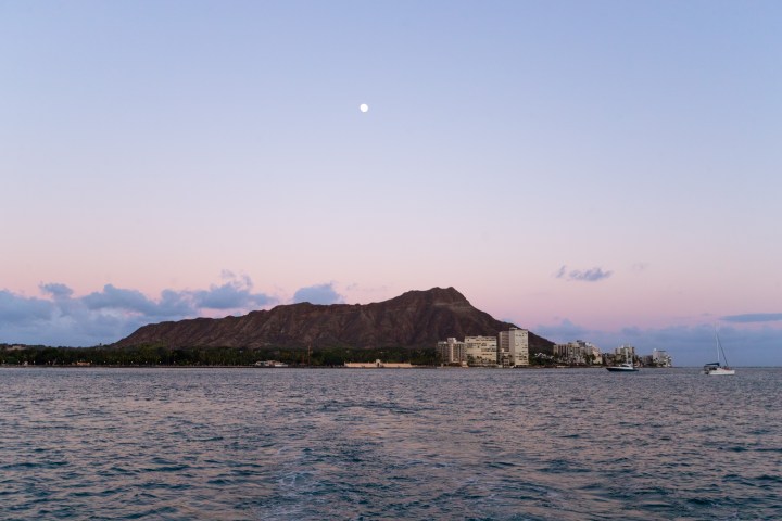 Coastal cityscape with mountains and moon under a pastel sky at sunset.