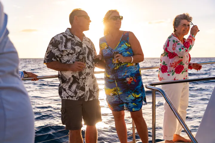 Three people on a boat at sunset, wearing floral shirts, enjoying a drink and the ocean view.