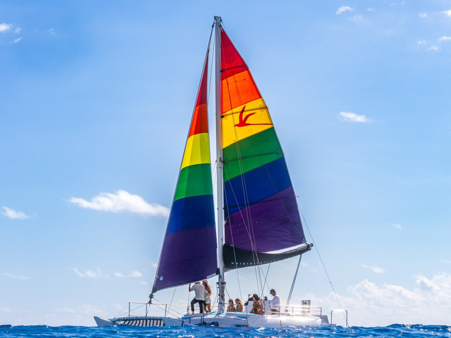 Sailboat with rainbow sails on blue ocean under clear sky with people on deck.