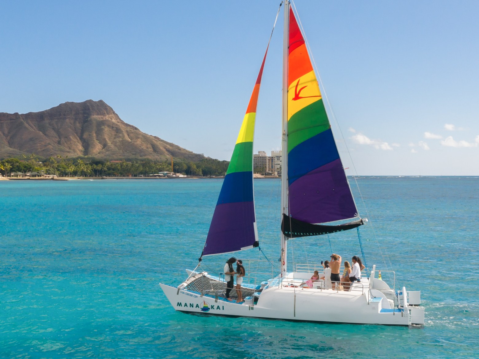 Sailboat with rainbow sail on blue ocean, mountain in background.