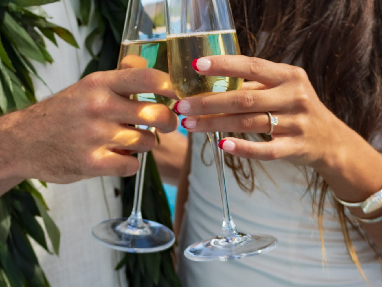 Two people clinking champagne glasses, one wearing a leafy garland, the other with pink manicured nails and ring.