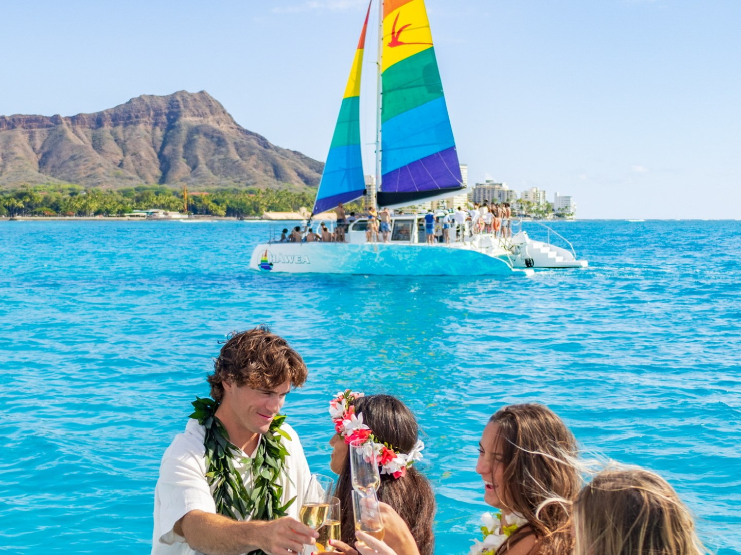 Group celebrating on a boat with leis, near a rainbow-sailed catamaran and scenic coastline in the background.