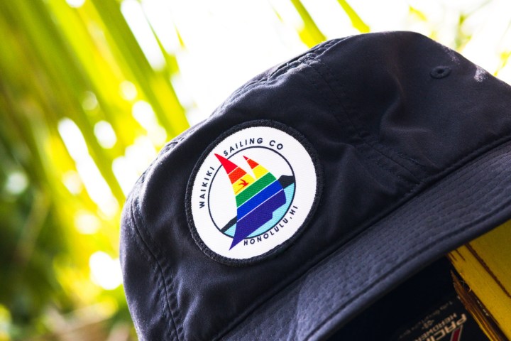 Black bucket hat with rainbow sailboat patch, palm leaves in background.